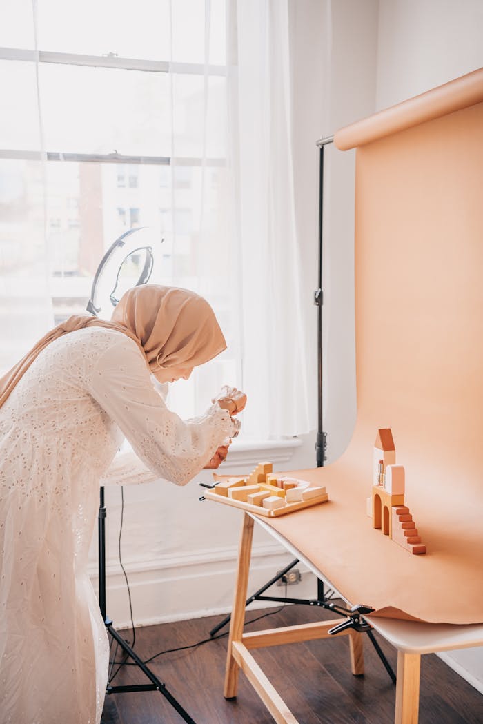 A young Muslim woman with a hijab photographing a miniature setup in a sunlit studio.